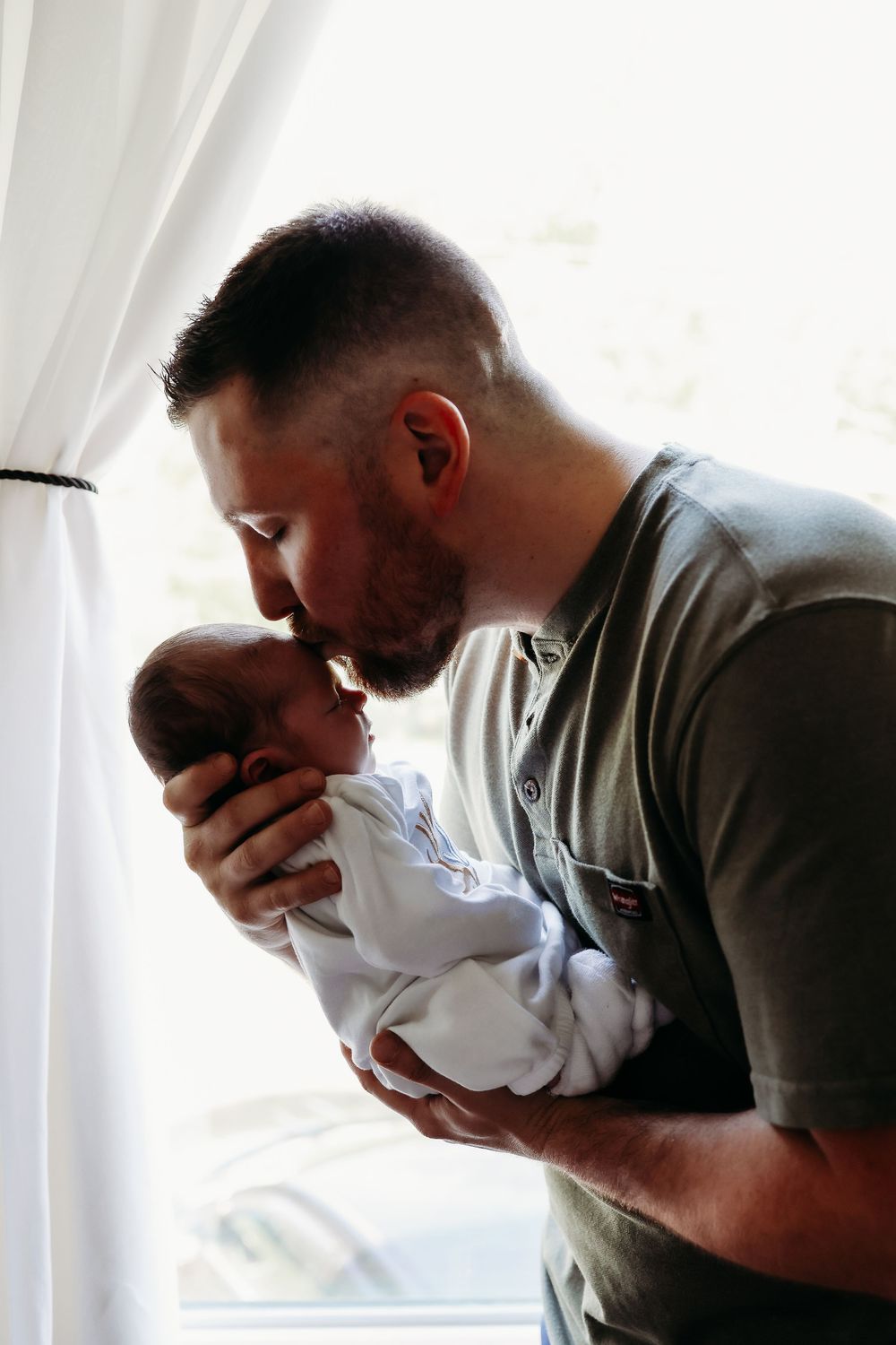 A man in a grey t-shirt holds and kisses a newborn baby near a window in a series of tender moments.
