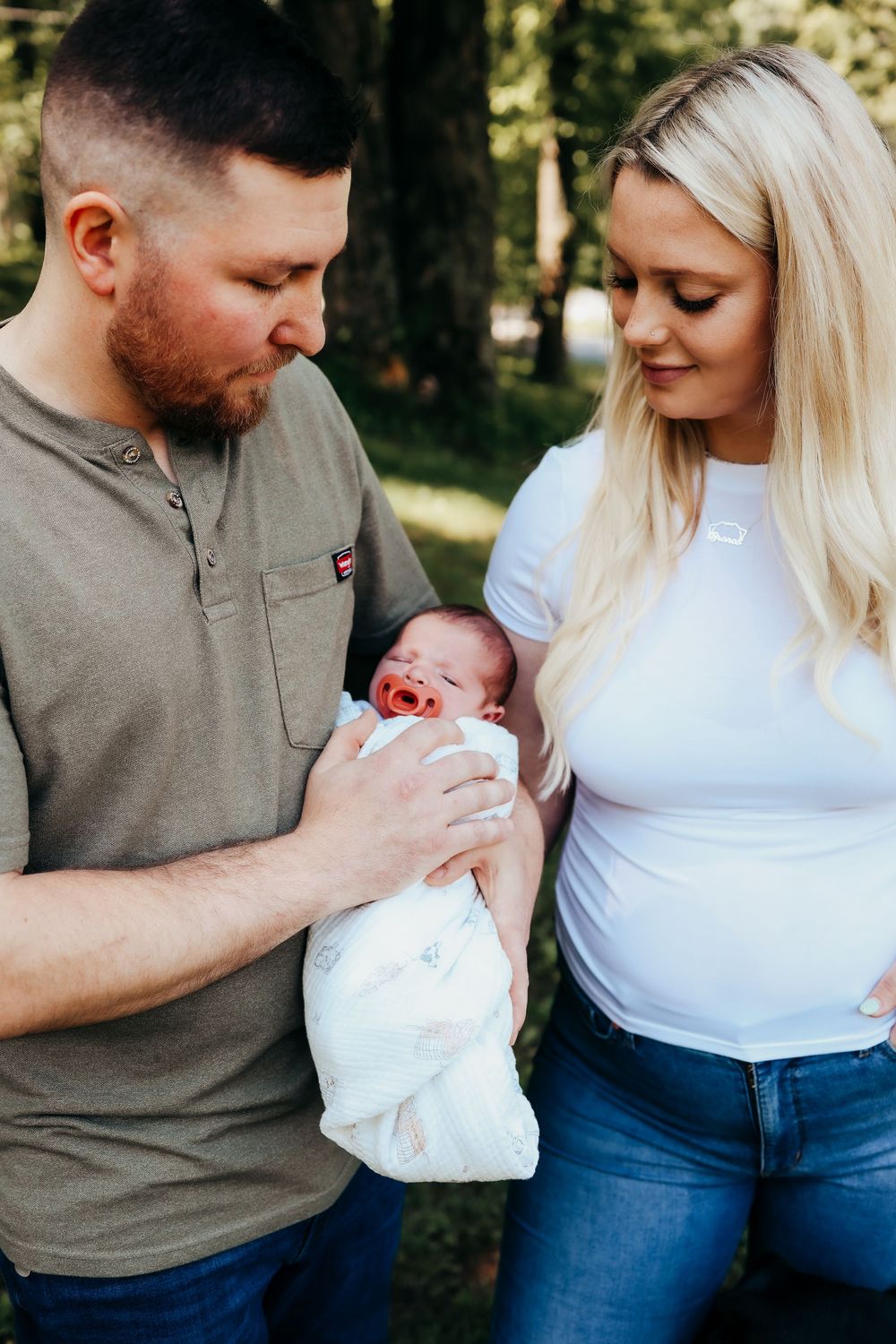 A couple outdoors gazes lovingly at their newborn baby wrapped in a white blanket.