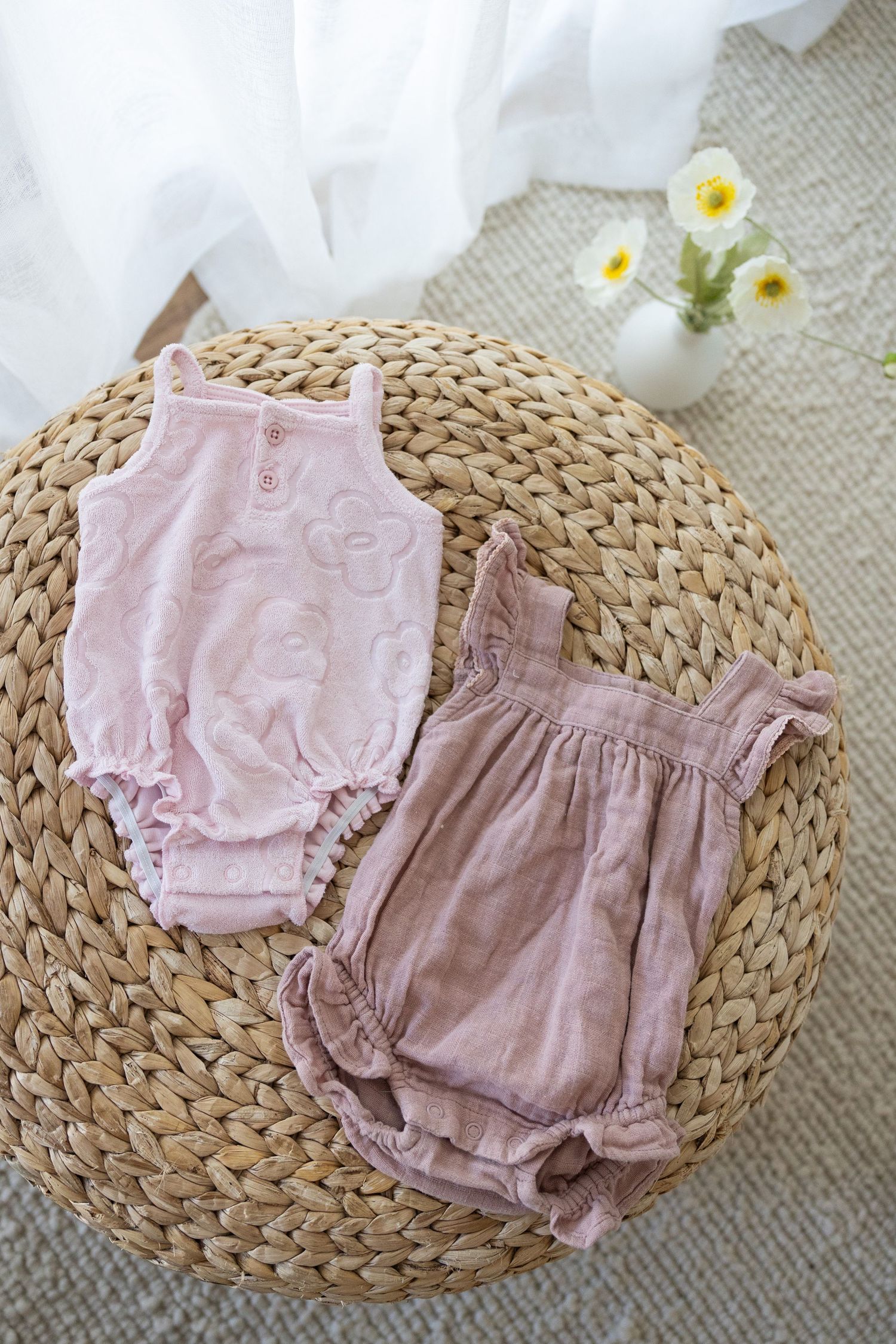 Two soft pink baby rompers displayed on a round woven rattan mat with delicate white flowers in a vase.
