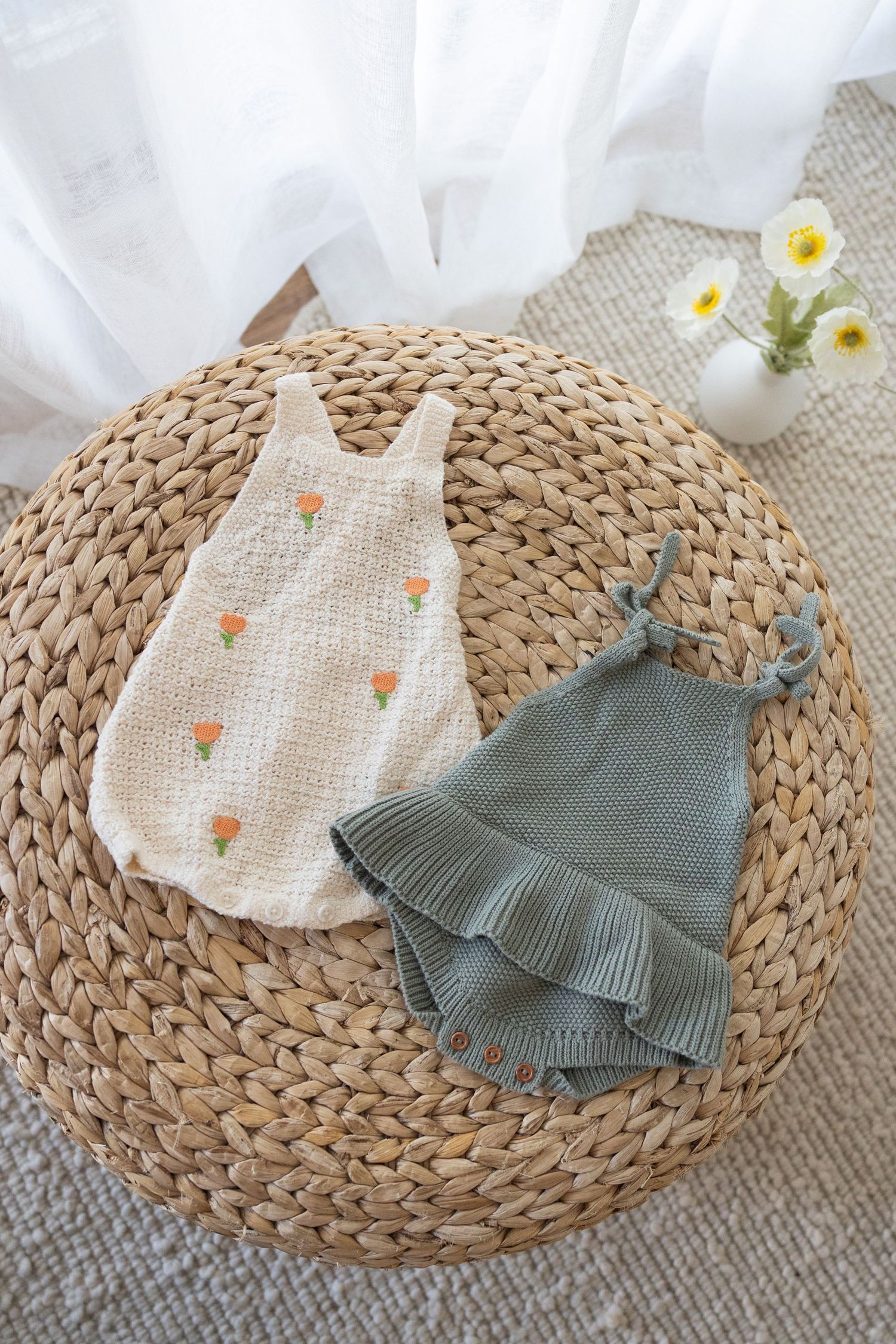 Two baby rompers displayed on woven rattan mat, one cream with orange flowers and one sage green with ruffles.