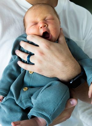 Newborn in blue outfit yawning while being held by adult hand with ring.