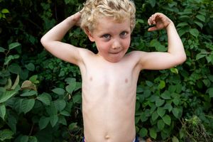 Young child flexing muscles shirtless against green foliage background.