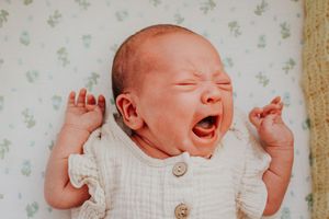 Newborn baby in white outfit yawning while lying on floral patterned background.