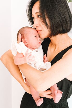 A tender moment between mother and newborn baby in white against light background.