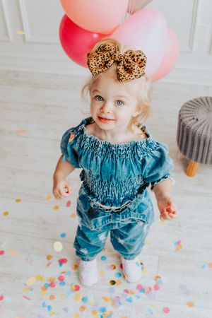 Toddler in ruffled teal outfit playing with pink balloons and confetti on white floor.