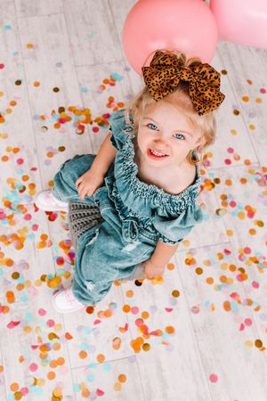 Happy toddler in blue ruffled dress lying on floor with colorful confetti and pink balloons.