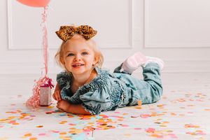 Child in denim romper and orange bow playing with confetti on white floor.