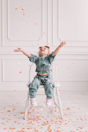 Joyful child in blue outfit throwing confetti while sitting on white chair.