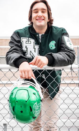Player wearing green helmet leans against chain link fence with athletic gear.