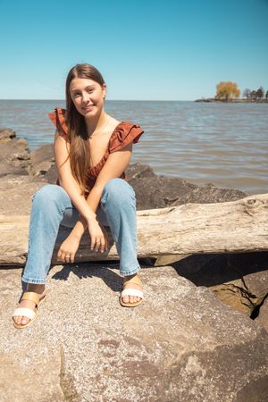 A casual summer portrait sitting on rocks by a lakeside wearing denim and a rust-colored top.