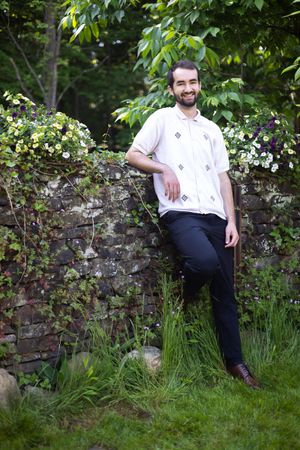 Person in white shirt poses casually against stone wall surrounded by lush greenery.