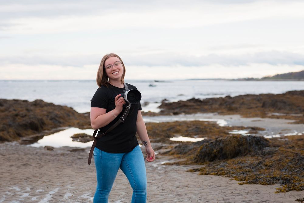 A beach-goer holds a small black puppy while standing on a rocky coastal shore at dusk.