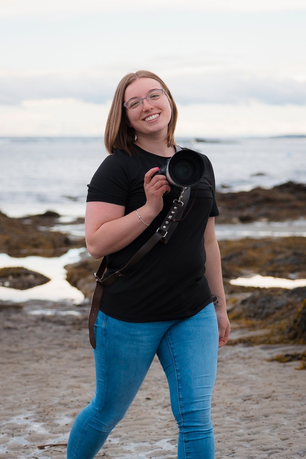 Person in black t-shirt and blue jeans standing on rocky beach with ocean and cloudy sky in background.