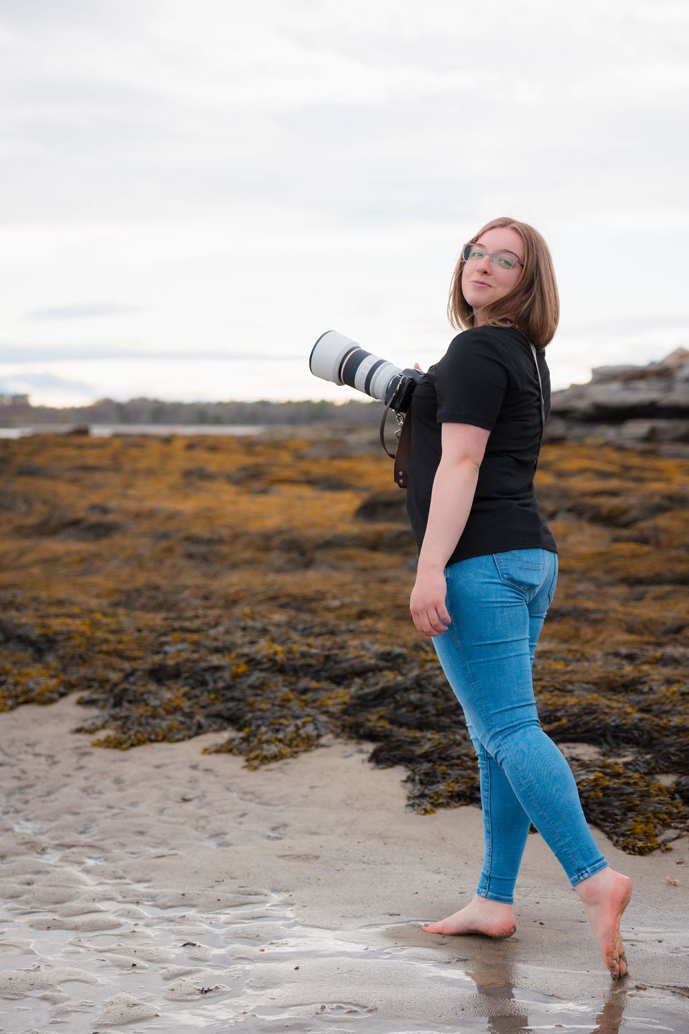Photographer with telephoto lens poses barefoot on a beach near rocky coast with autumn colored vegetation.