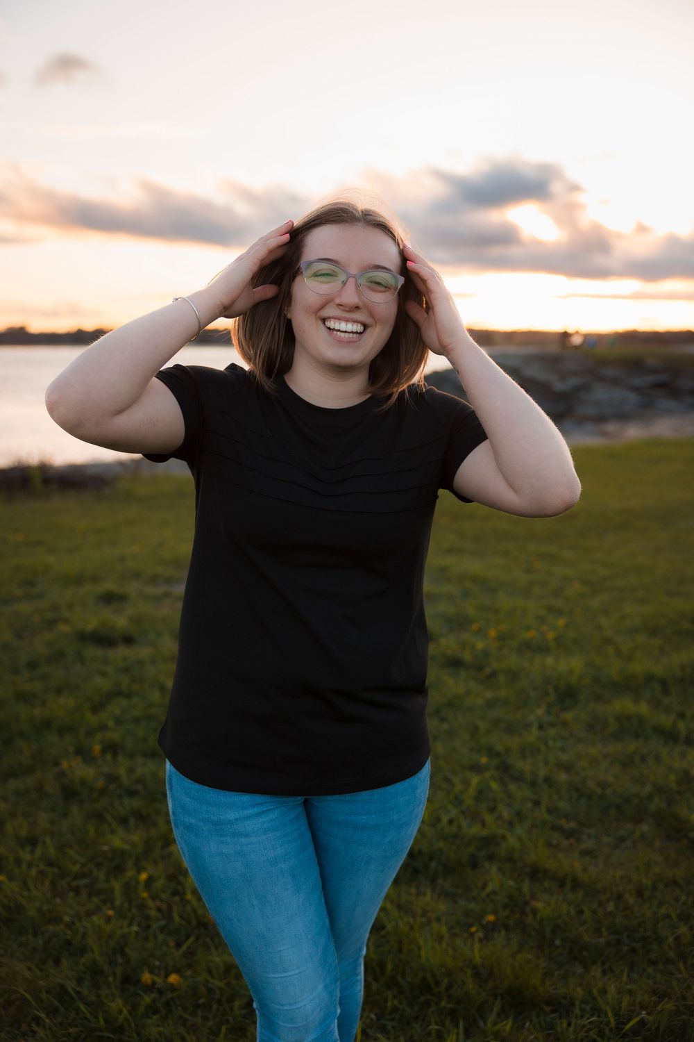 Person wearing glasses and black t-shirt smiles joyfully at sunset in a grassy outdoor setting by the water.