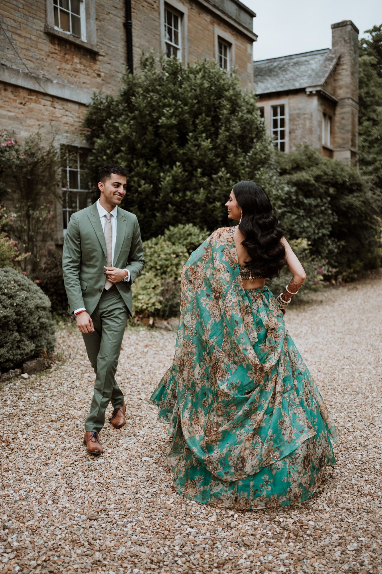 A couple walks on a gravel path outside a rustic stone building, wearing green formal attire and a flowing floral gown.