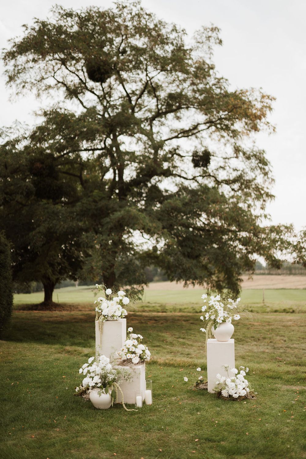 White floral wedding decorations with tiered displays placed on grass under a majestic oak tree in a rustic outdoor setting.