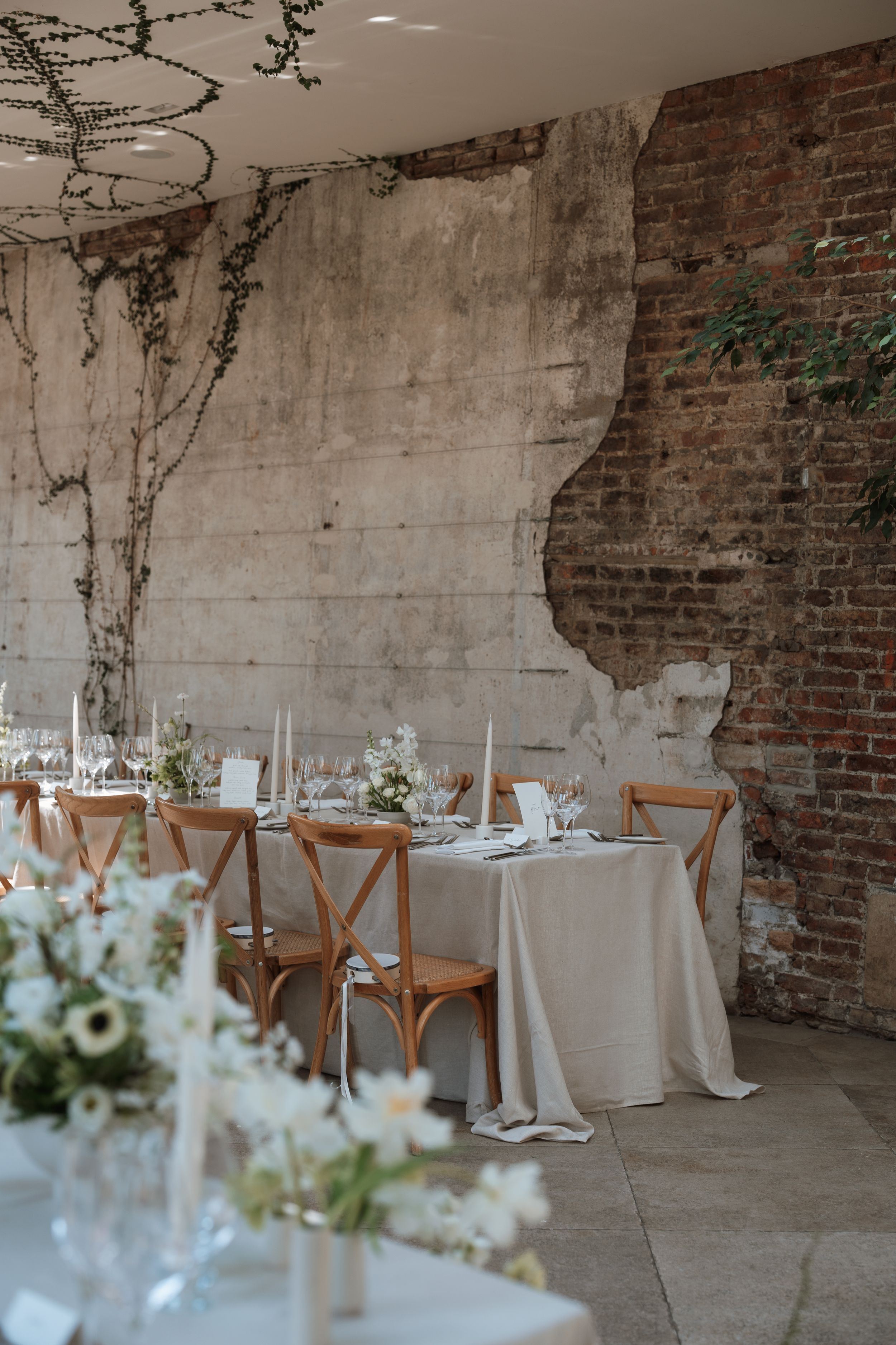 Rustic wedding reception setup with white table linens, wooden cross-back chairs and elegant floral centerpieces against brick wall.