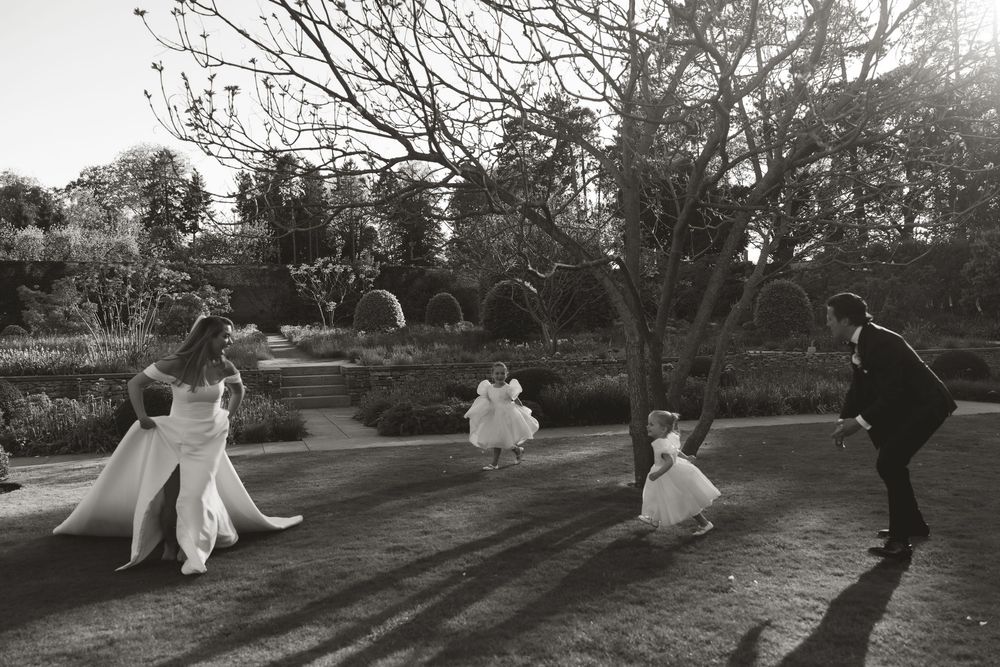A black and white photograph shows people dancing and playing together in a garden at sunset, casting long shadows.