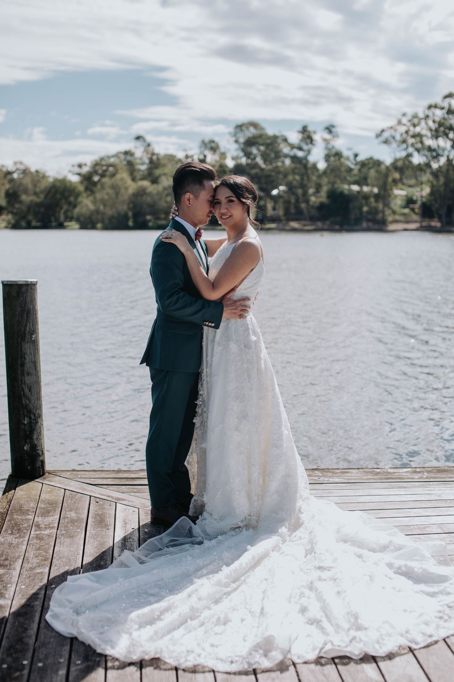 A couple embraces on a wooden dock by a tranquil lake, with a stunning white wedding gown trailing across the boards.