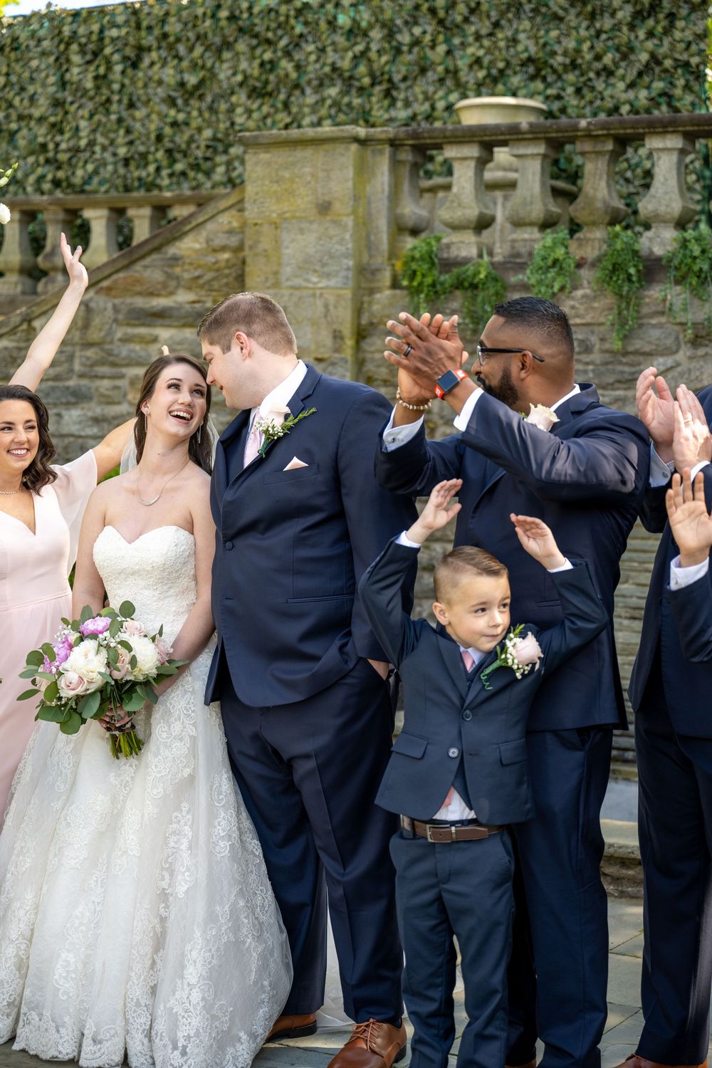 Wedding party celebrating newlyweds on stone steps, bride in lace gown holding bouquet, groomsmen in navy suits cheering.