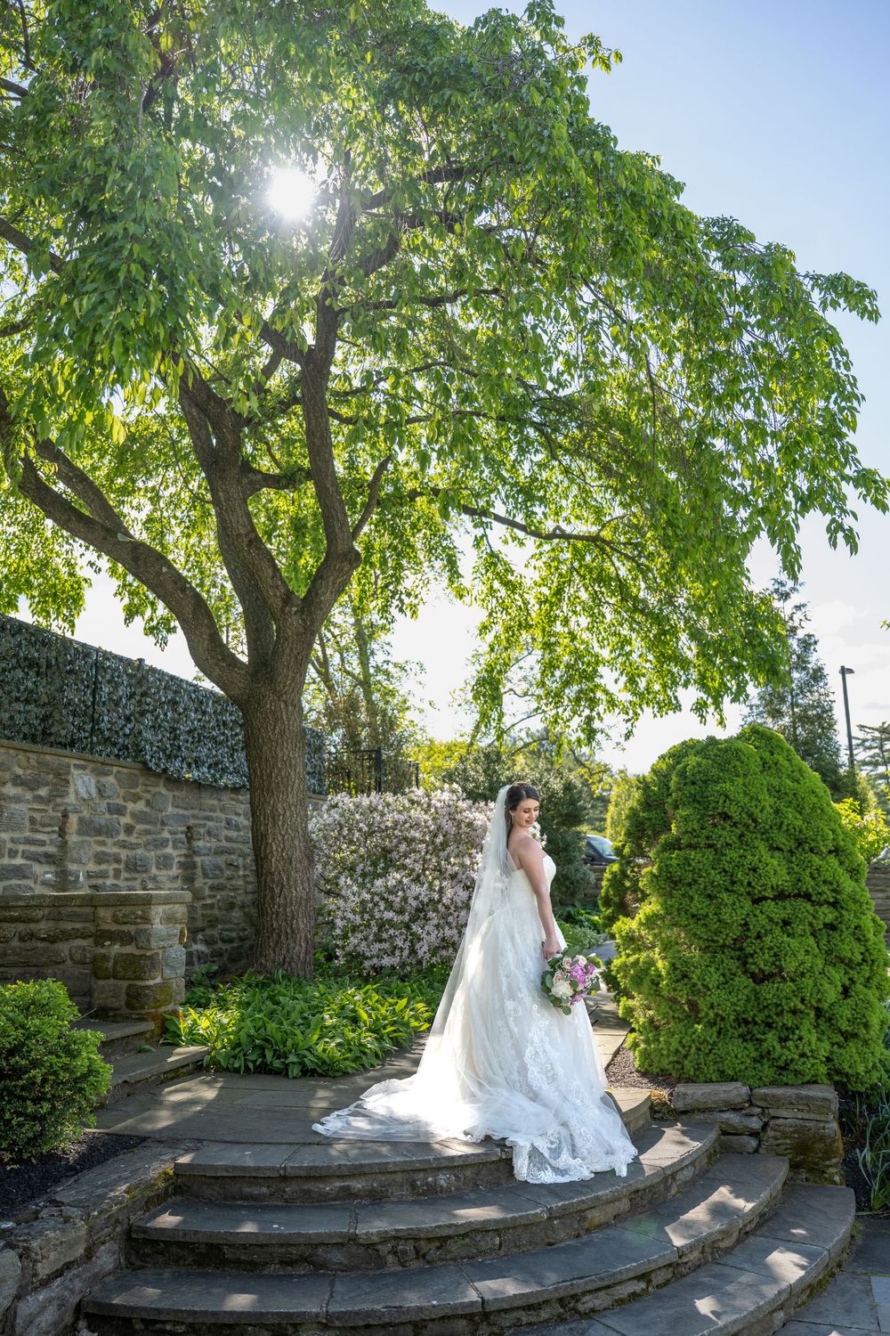 Bride in white lace gown holding bouquet stands on stone steps in lush garden with sunlight filtering through tree.
