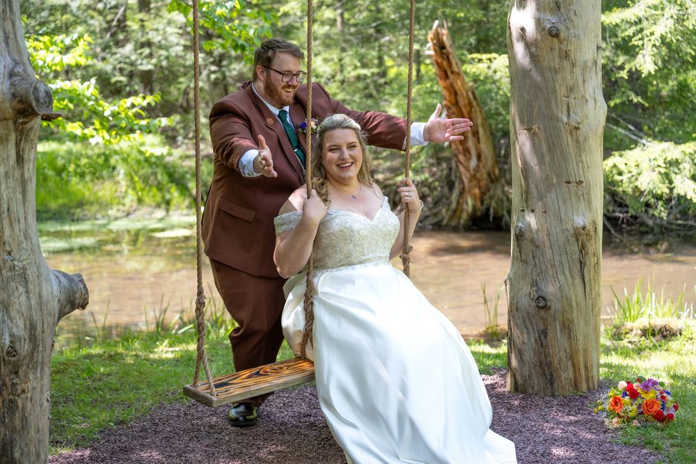 Bride on wooden swing in white gown, groom in brown suit pushing her, outdoor woodland wedding setting.