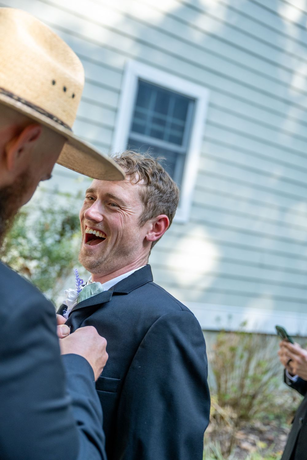 A laughing groomsman in a dark suit gets his boutonniere adjusted outdoors near a white clapboard house.