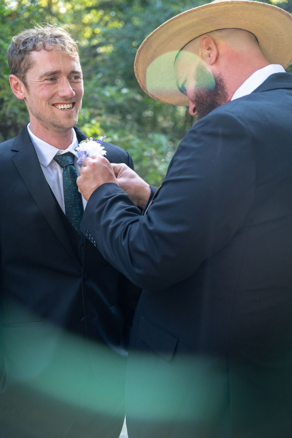 A smiling groom in a dark suit receives a boutonniere from a man in a straw hat at an outdoor wedding.