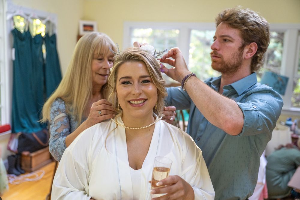 Bride smiling as hairstylist and helper adjust her hair accessory while she holds a champagne glass.