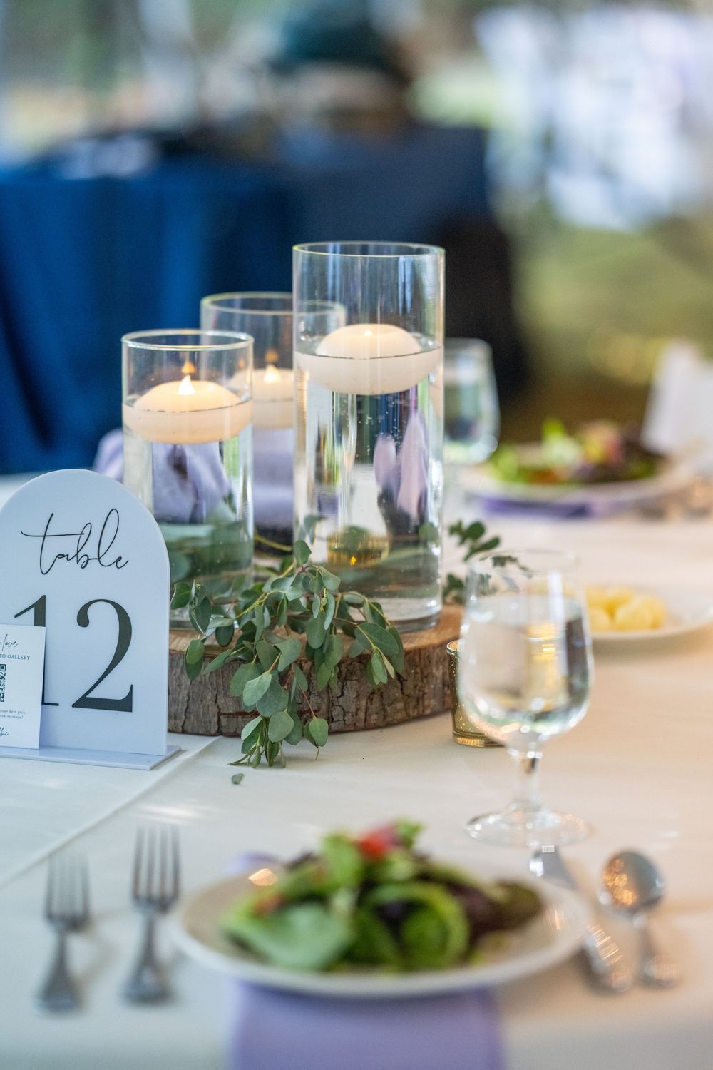 Elegant wedding reception table 12 with floating candles in glass cylinders, greenery on wood slice, and fresh salad plates.