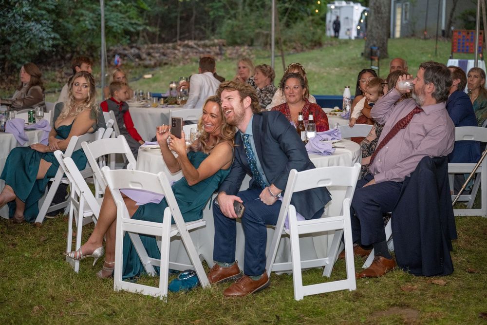 Guests laughing and taking photos at an outdoor wedding reception seated at white round tables.