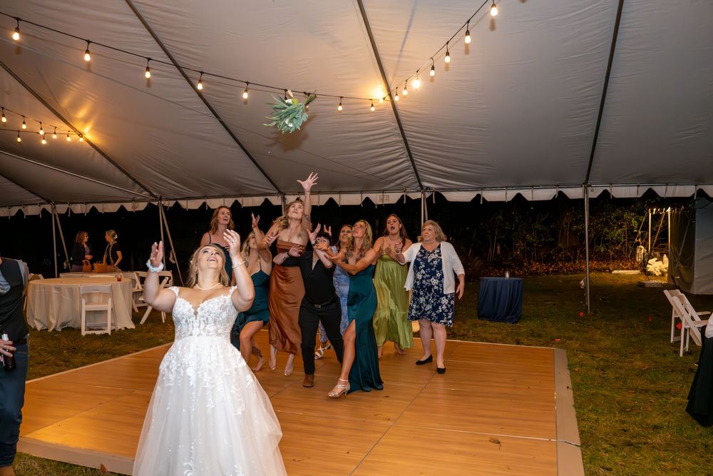 Bride tossing bouquet to excited guests on dance floor under a white tent at wedding reception.