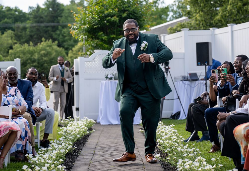 A joyful groom in a green suit dances down the wedding aisle at an outdoor ceremony with seated guests.