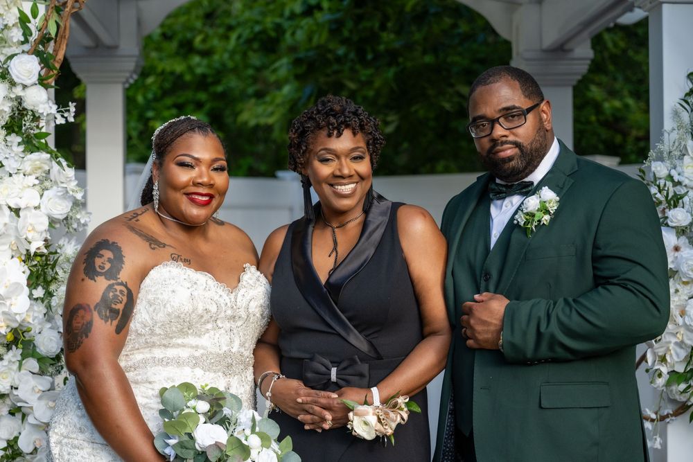 Bride and groom in wedding attire pose with officiant outdoors near floral arch at elegant ceremony.