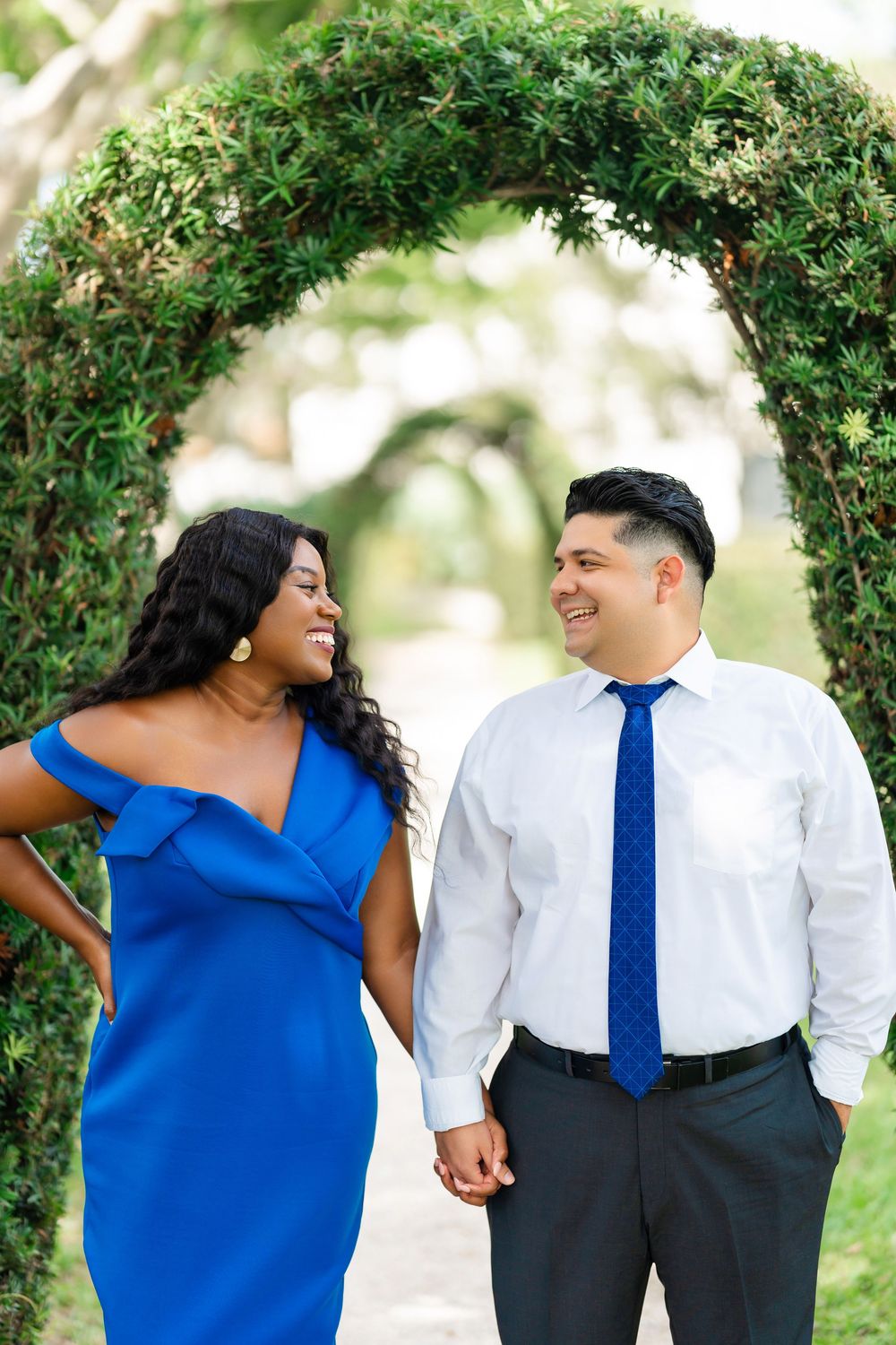 Couple standing together under lush green garden archway during engagement photoshoot, with woman in blue dress and man in tie.