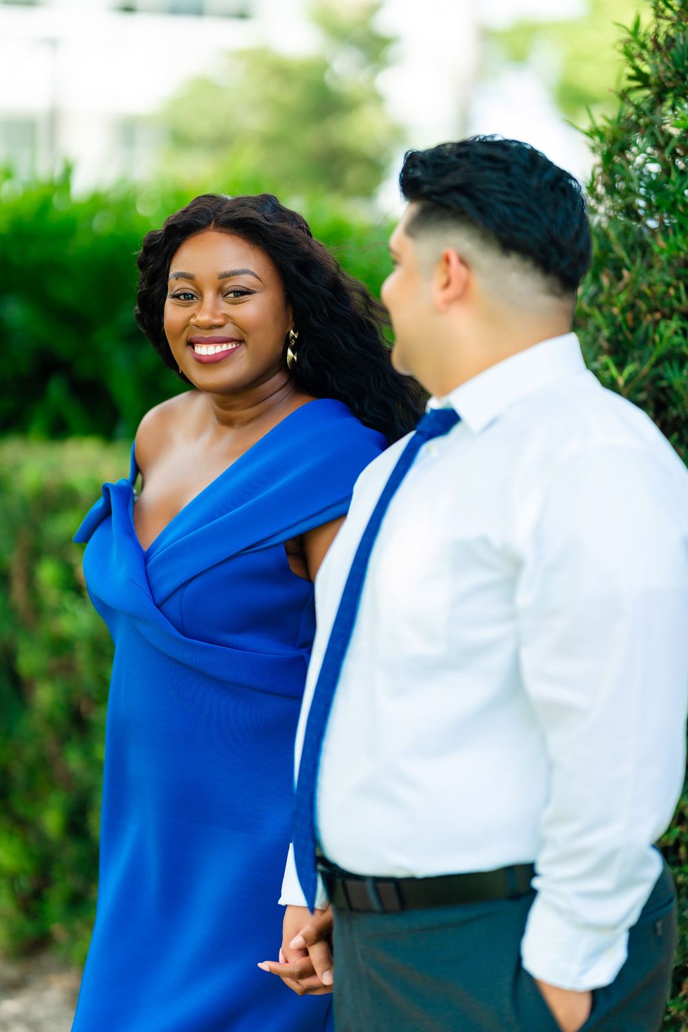 A couple in formal attire, with a royal blue dress and white dress shirt with suspenders, share a moment in a garden setting.