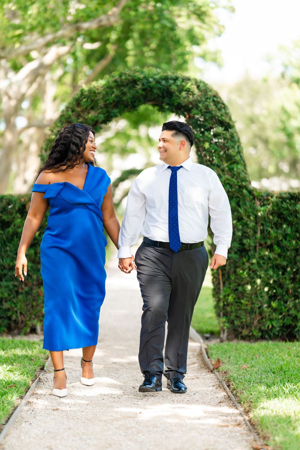 A couple in coordinated blue outfits walks together holding hands under a leafy garden archway.
