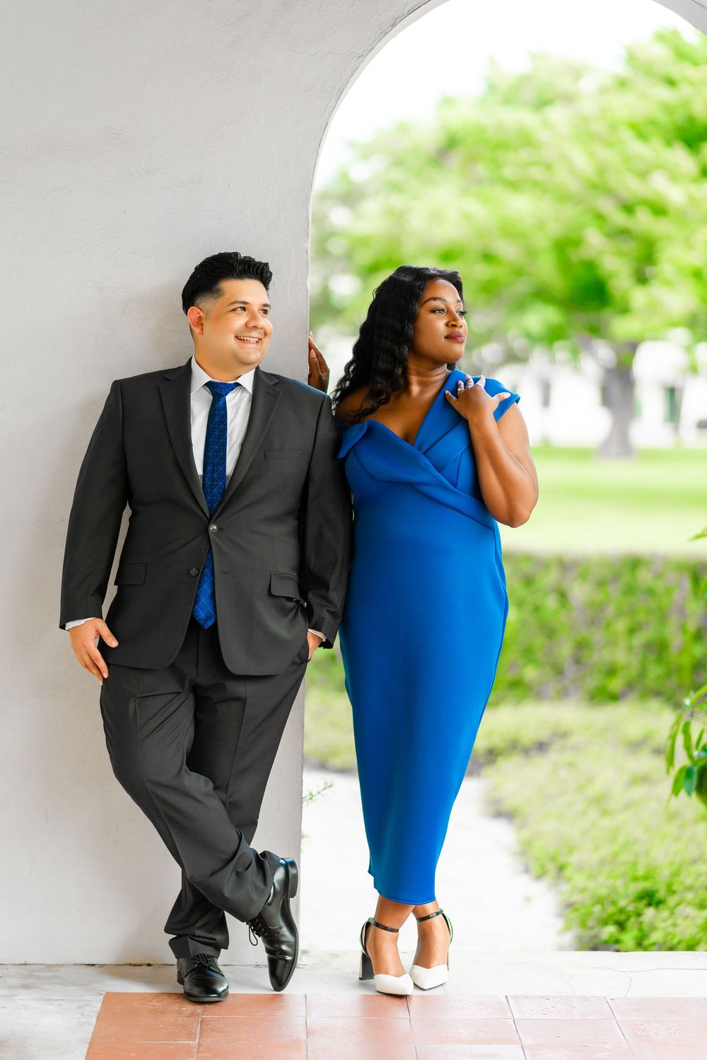 A couple poses elegantly against a white archway, with one wearing a dark suit and the other in a blue dress.