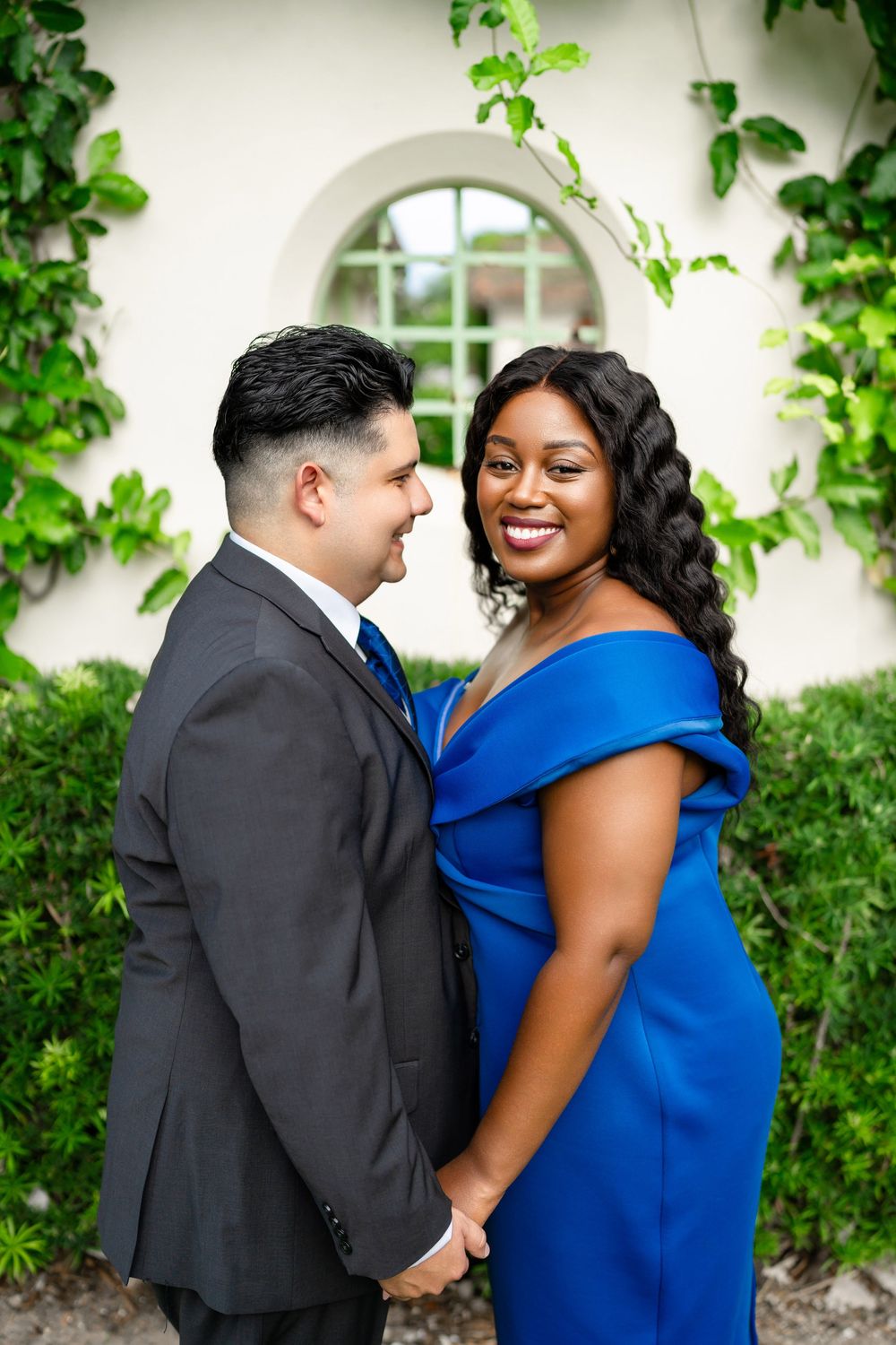 A couple shares a romantic moment in front of an arched doorway draped with green ivy, wearing formal attire.