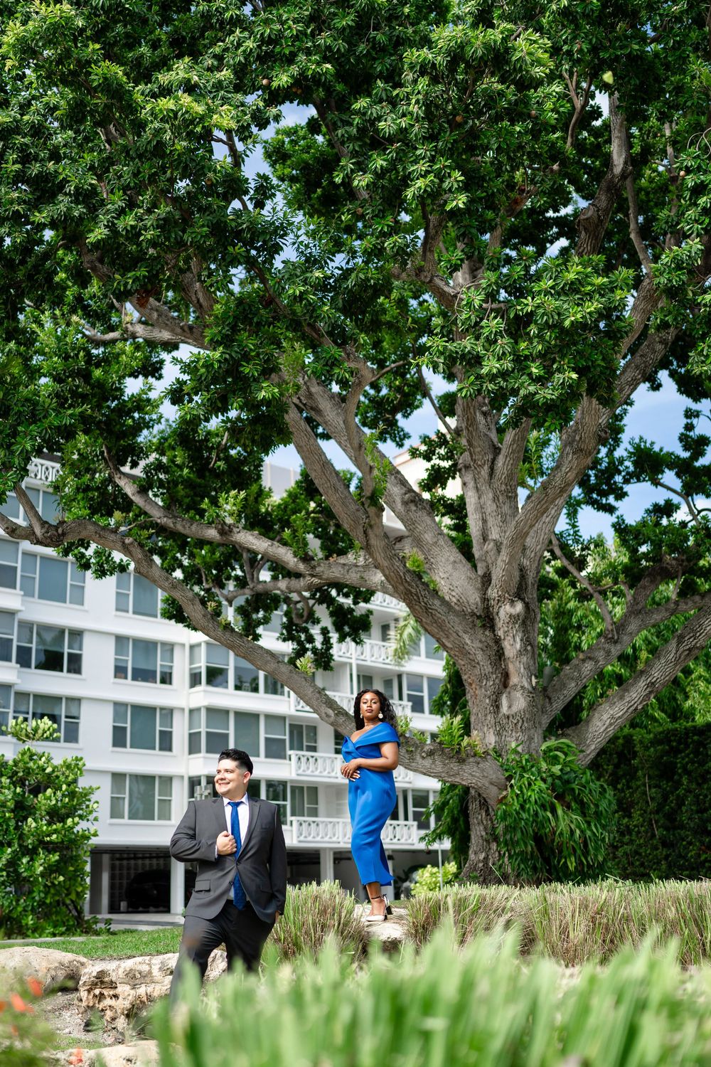 A couple stands near a large leafy tree outside a modern building with one wearing a blue dress and another in business attire.