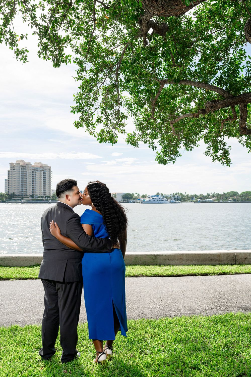 A couple embraces by the waterfront with city buildings and a large tree in the background.