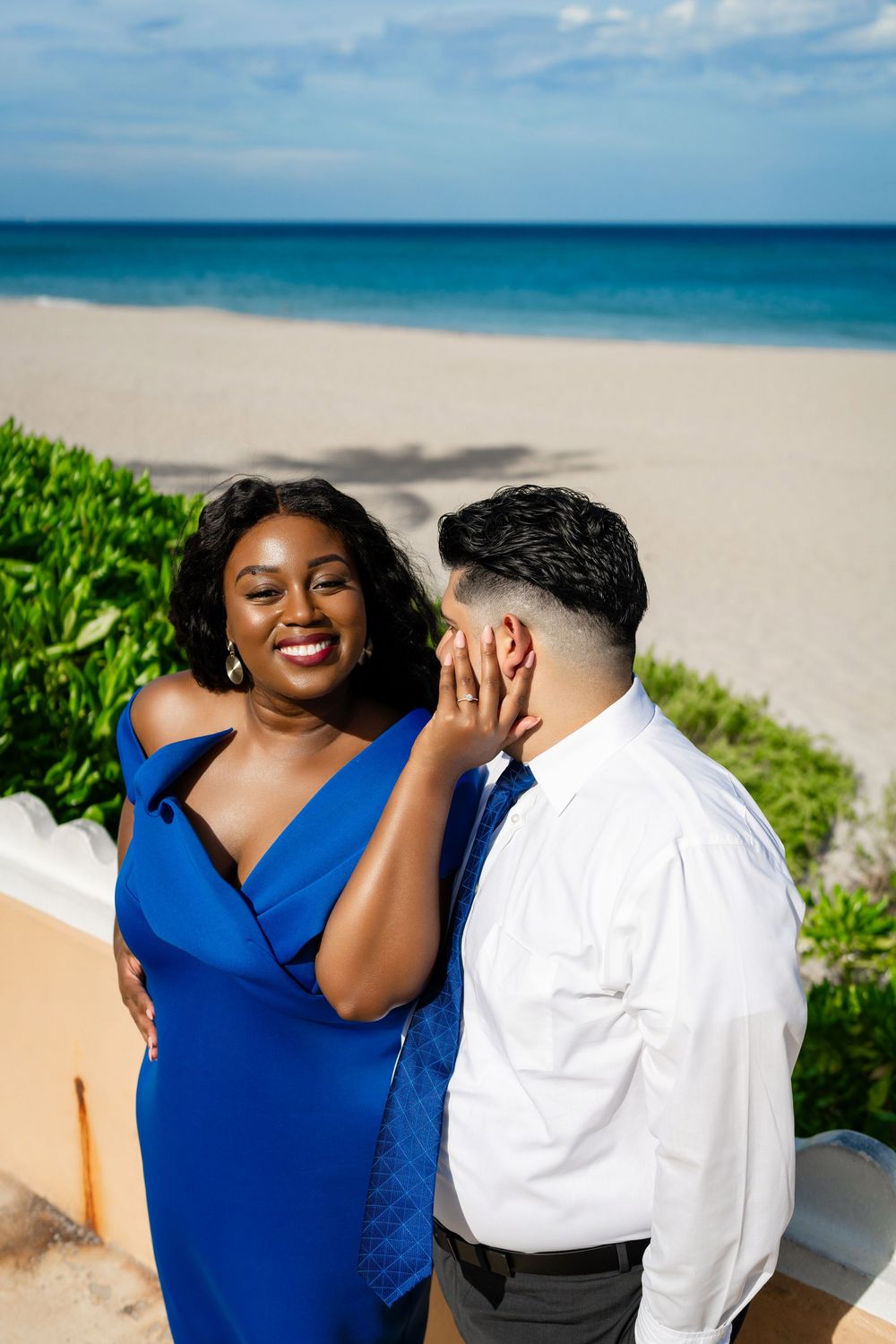 A couple shares a joyful moment on a tropical beach with turquoise waters and green foliage in the background.