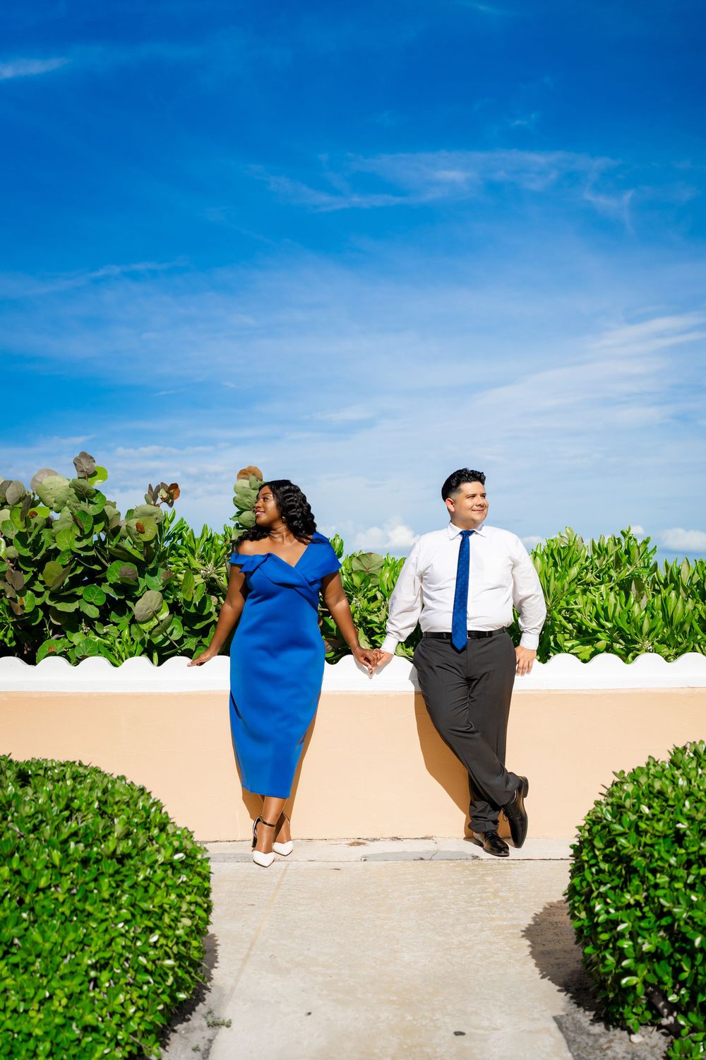 A couple in blue dress and business attire pose against a wall with lush greenery and bright blue sky.