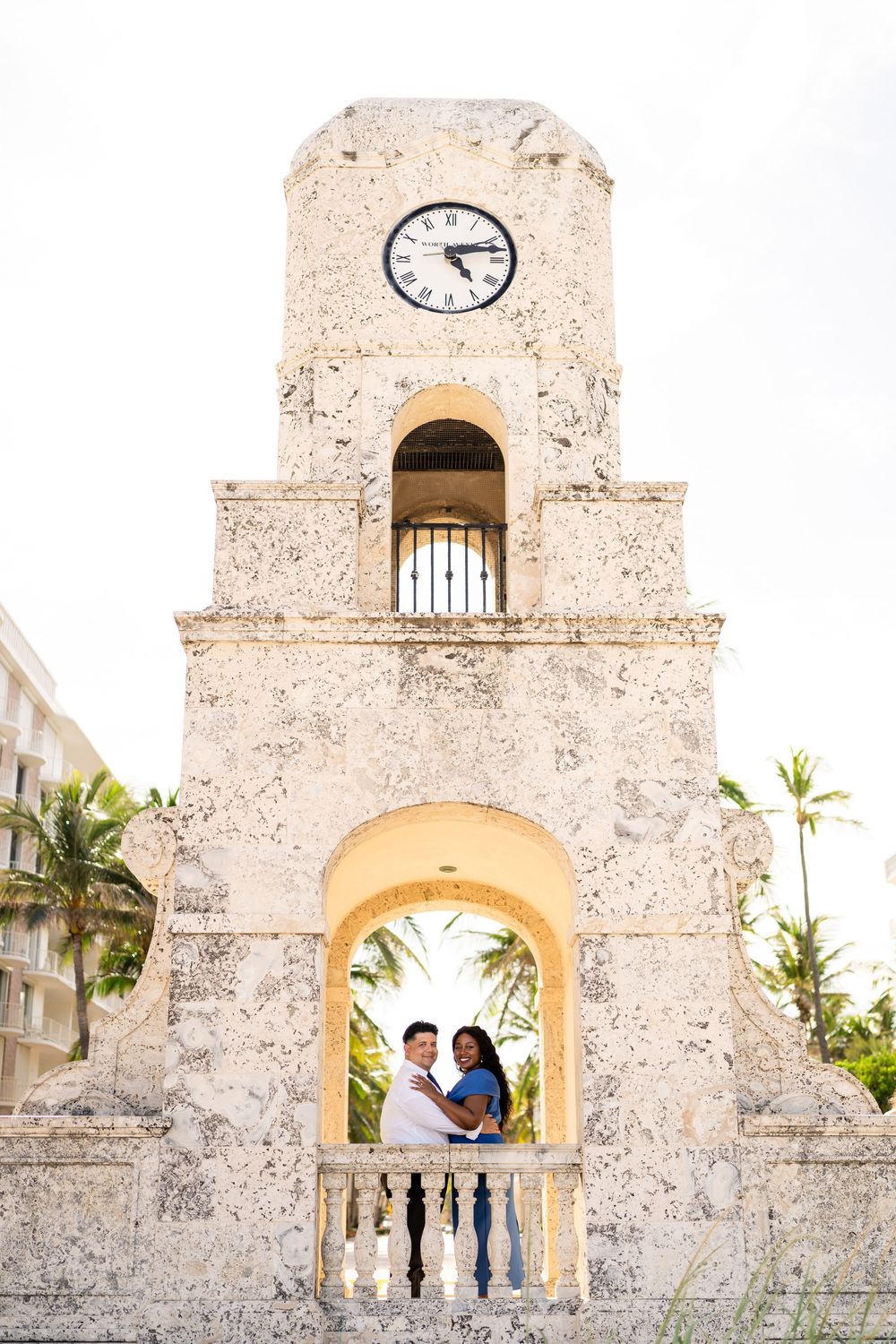 A couple poses in an archway of a historic clock tower with palm trees visible.