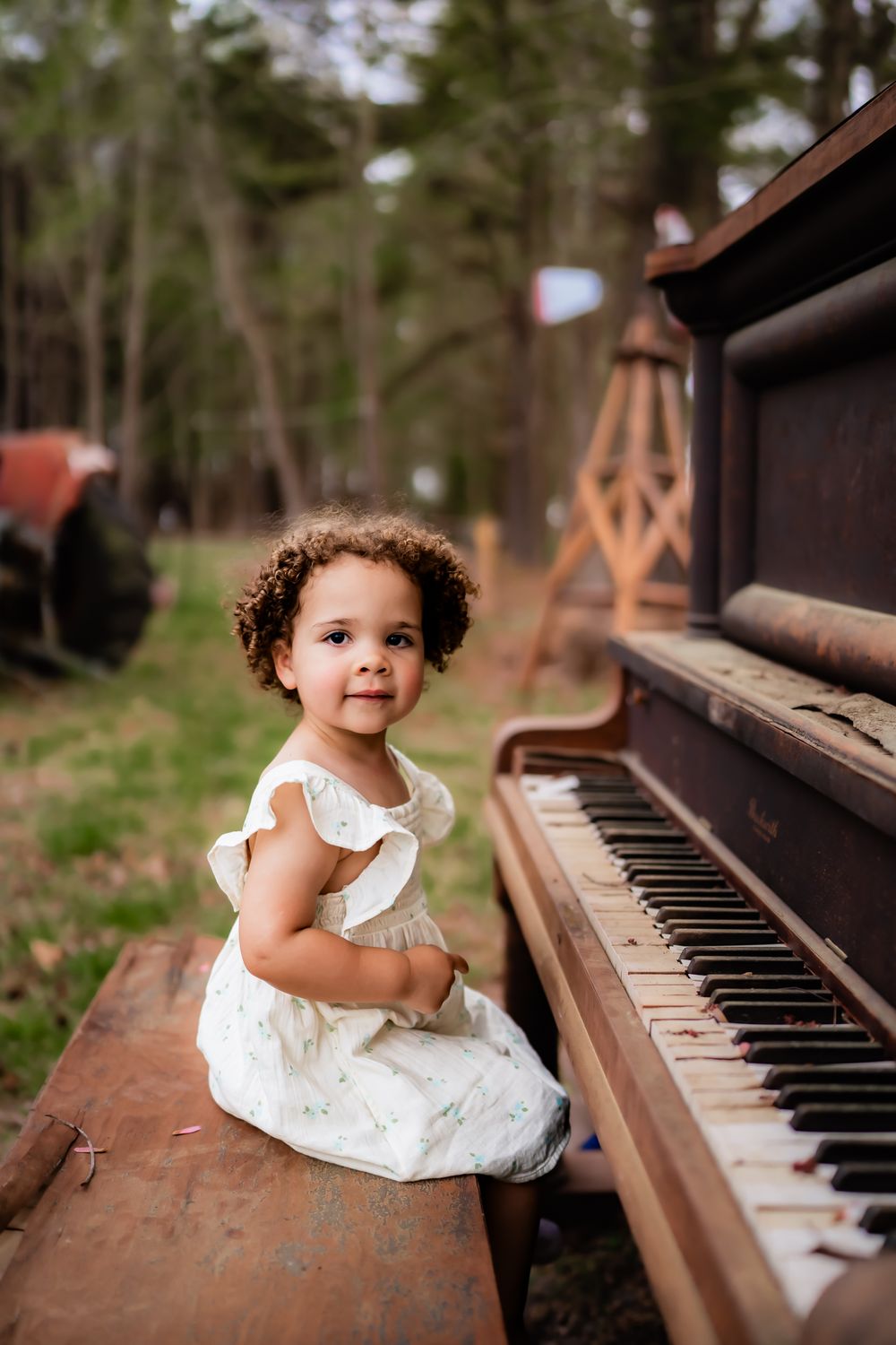 A small child in a white dress sits at an outdoor piano amidst a natural wooded setting.