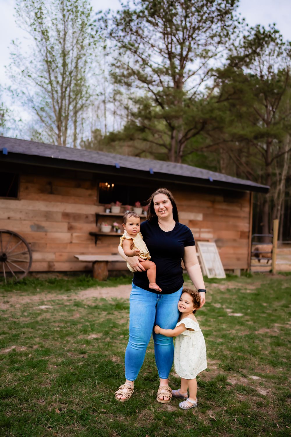 A woman in a black shirt and jeans stands outside a rustic wooden cabin with two small children in a grassy yard.