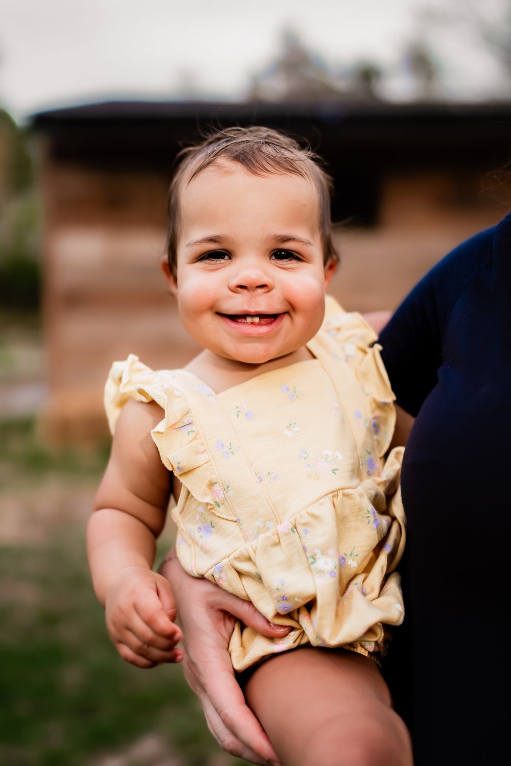 Sweet toddler wearing a pale yellow ruffled romper smiles brightly while being held outdoors.