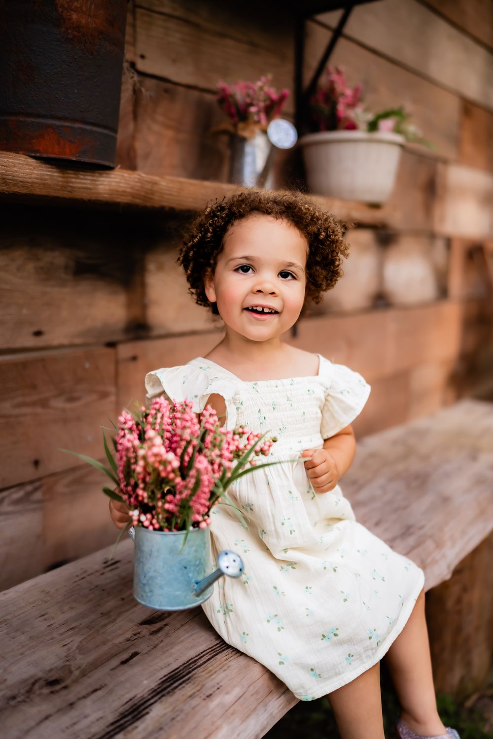 Child in white eyelet dress sits on rustic wooden steps next to blue vase of pink flowers.