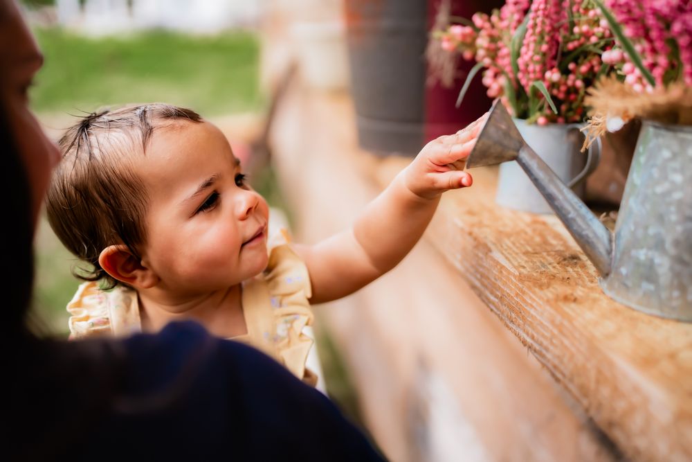 Small hand reaches up to touch delicate pink and purple flowers in a rustic vase on wooden surface.